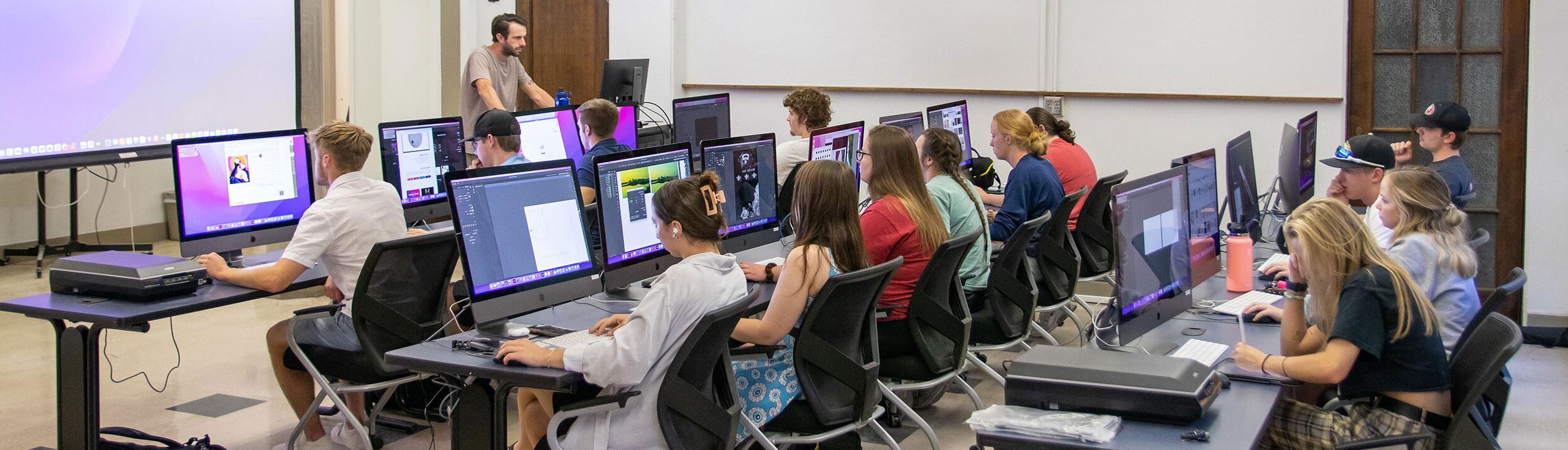 Classroom with three rows of desks with computers with big monitors. Students are seated at desks working with an instructor at the front of the class.
